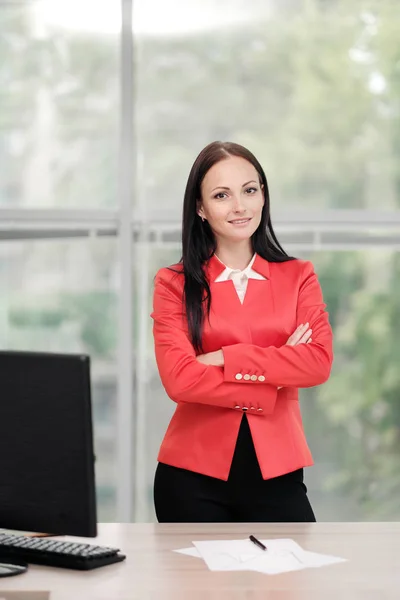 En ung attraktiv hvid blond kvinde i rødt jakkesæt sidder ved et skrivebord på et lyst kontor. Portræt af en forretningsmand, direktør for organisationen . - Stock-foto