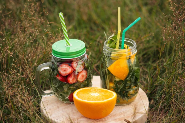 Dois copos de limonada com túbulos estão em uma placa de madeira na grama verde. Bebidas de verão de morangos e laranjas com hortelã e alecrim . — Fotografia de Stock
