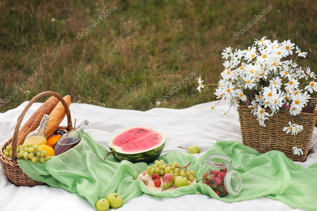 Summer picnic in the meadow on the green grass. Fruit basket, juice and bottled wine, watermelon and bread baguettes. White tablecloth and a bouquet of field daisies.