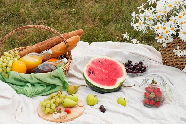 Summer picnic in the meadow on the green grass. Fruit basket, juice and bottled wine, watermelon and bread baguettes. White tablecloth and a bouquet of field daisies.