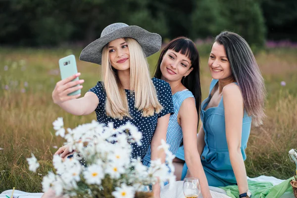 Três mulheres jovens, em vestidos azuis e chapéus sentam-se em uma xadrez e tiram fotos em um smartphone. Piquenique ao ar livre na grama na floresta. Comida deliciosa em uma cesta de piquenique e vinho . — Fotografia de Stock