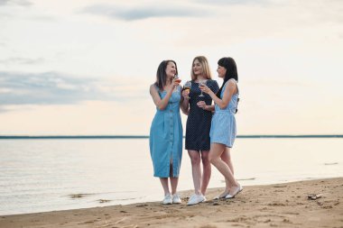 Üç genç çekici kadın, mavi elbiseler deniz kıyısında yürüyor. Kız iletişim, gülmek ve şarap içmek.