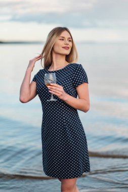 Genç ve çekici bir sarışın kadın, mavi bir elbiseyle, sahil boyunca yürür. Bir bardak şarap ile tatile gülümseyen bir kadının portresi.