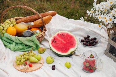 Yeşil çim üzerinde çayırda yaz pikniği. Meyve sepeti, meyve suyu ve şişe şarabı, karpuz ve ekmek bagetleri. Beyaz masa örtüsü ve bir buket alan papatyalar.