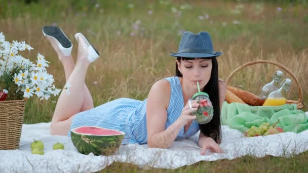 Una joven hermosa mujer en un sombrero y vestido está bebiendo limonada de una lata mientras está sentada en un cuadros en la hierba verde. Cesta de picnic, un ramo de margaritas, sandía . — Vídeos de Stock