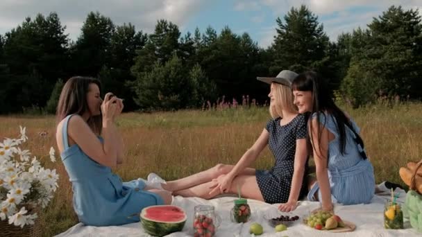 Tre giovani ragazze attraenti su un picnic. Il fotografo scatta foto sulla fotocamera mirrorless di due modelli. Modelli posare e guardare le foto . — Video Stock
