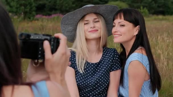 피크닉에 세 젊은 매력적인 여자. 사진작가는 두 모델의 미러리스 카메라로 사진을 찍습니다. 모델 포즈 및 사진 보기. — 비디오