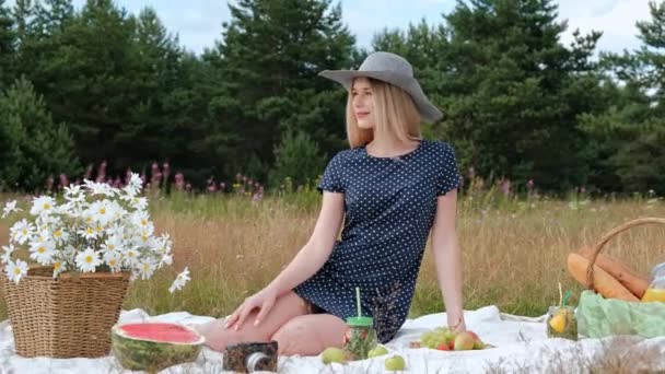 En ung vacker blond kvinna i en hatt och klänning dricker lemonad från en kan medan du sitter på en pläd på det gröna gräset. Picknickkorg, en bukett av prästkragar, vattenmelon. — Stockvideo