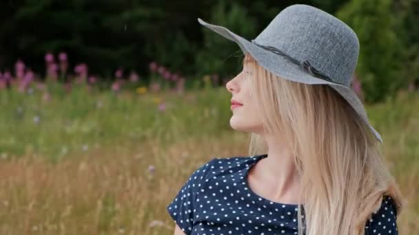 Una joven mujer rubia atractiva con un vestido azul y un sombrero está sentada sobre una tela a cuadros en la hierba. El viento sopla pelo . — Vídeos de Stock