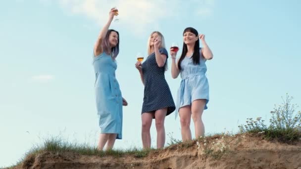 Tre giovani donne attraenti, in abiti blu stanno camminando lungo la riva del mare. Le amiche comunicano, ridono e bevono vino . — Video Stock