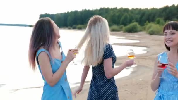 Drei junge attraktive Frauen in blauen Kleidern spazieren an der Küste entlang. Freundinnen kommunizieren, lachen und trinken Wein. — Stockvideo