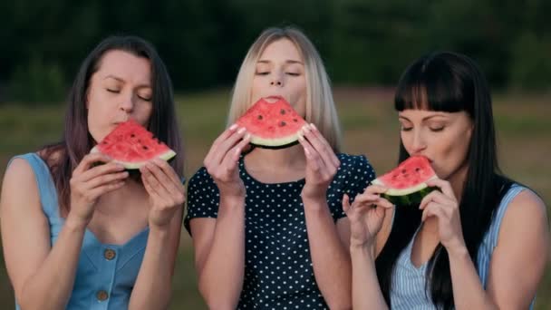 Tre unga attraktiva kvinnliga vänner i blå klänningar vid solnedgången äter vattenmelon och leende. — Stockvideo