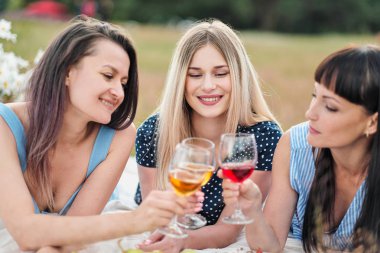 Mavi elbiseler içinde üç genç kadın, ve şapka ekose ve İçecek şarap üzerinde yatıyor. Sahilde çim üzerinde açık piknik. Piknik sepeti ve şarap lezzetli yemek. Karpuz, üzüm ve buket.