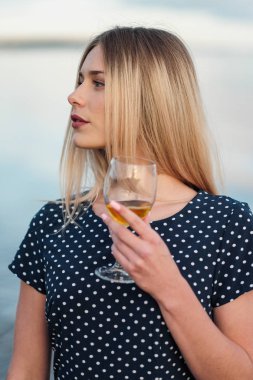 Genç ve çekici bir sarışın kadın, mavi bir elbiseyle, sahil boyunca yürür. Bir bardak şarap ile tatile gülümseyen bir kadının portresi.