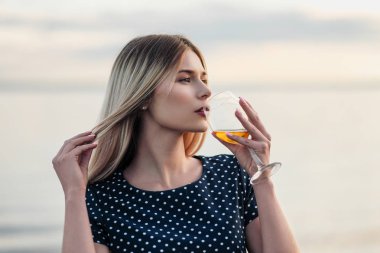 Genç ve çekici bir sarışın kadın, mavi bir elbiseyle, sahil boyunca yürür. Bir bardak şarap ile tatile gülümseyen bir kadının portresi.