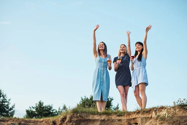 青いドレスを着た3人の若い魅力的な女性が海岸沿りを歩いています。ガールフレンドがワインをコミュニケーション、笑い、飲む. — ストック写真
