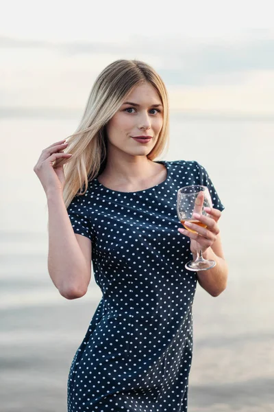 Młoda atrakcyjna blond kobieta, w niebieskiej sukni, spacery wzdłuż brzegu morza. Portret uśmiechniętej kobiety na wakacjach z kielichem wina. — Zdjęcie stockowe