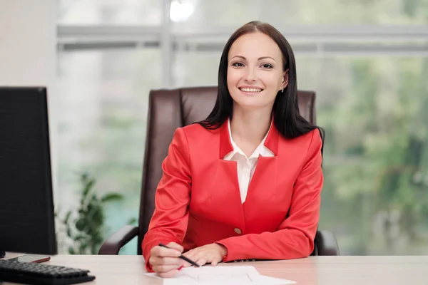 Una joven y atractiva rubia caucásica vestida con un traje de negocios rojo se sienta en un escritorio en una oficina luminosa. Retrato de un empresario, director de la organización . — Foto de Stock