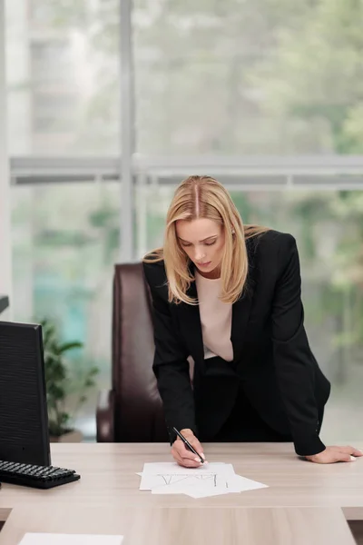 Jonge aantrekkelijke blanke blonde vrouw in zwart Business Suit zit aan de balie in Bright Office. Het bestuderen van papieren documenten, kijken naar camera en glimlachend. — Stockfoto