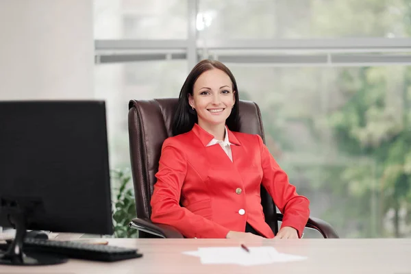 Una joven y atractiva rubia caucásica vestida con un traje de negocios rojo se sienta en un escritorio en una oficina luminosa. Retrato de un empresario, director de la organización . — Foto de Stock