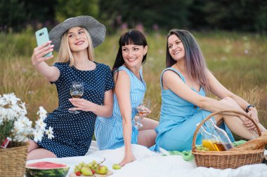 Üç genç kadın, mavi elbiseler ve şapkalar bir ekose üzerinde oturmak ve bir akıllı telefonda fotoğraf çekmek. Ormanda çim üzerinde açık piknik. Piknik sepeti ve şarabında lezzetli yemekler.