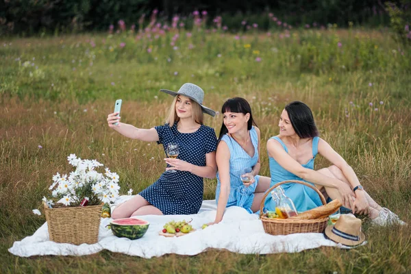 Três mulheres jovens, em vestidos azuis e chapéus sentam-se em uma xadrez e tiram fotos em um smartphone. Piquenique ao ar livre na grama na floresta. Comida deliciosa em uma cesta de piquenique e vinho . — Fotografia de Stock