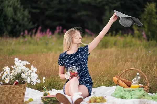 Young attractive woman in a blue dress at an outdoor picnic. A basket with daisies, watermelon, strawberries and a glass of lemonade.