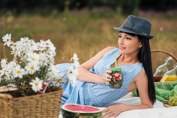 Молодая красивая женщина в шляпе и платье пьет лимонад из стеклянной банки, сидя на клетке на зеленой траве. Корзина для пикника, букет маргариток, арбуз . — стоковое фото
