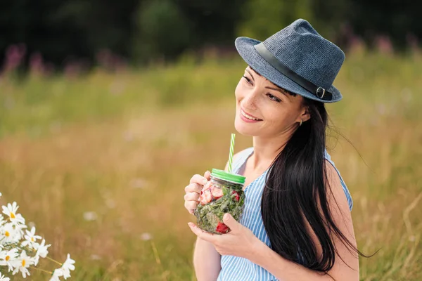 Une jeune belle femme en chapeau et robe boit de la limonade dans un bocal en verre, assise sur un plaid sur de l'herbe verte. Panier pique-nique, bouquet de marguerites, pastèque . — Photo