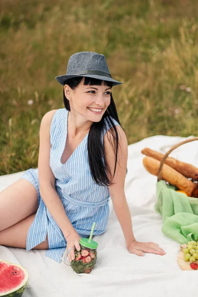 Молодая красивая женщина в шляпе и платье пьет лимонад из стеклянной банки, сидя на клетке на зеленой траве. Корзина для пикника, букет маргариток, арбуз . — стоковое фото