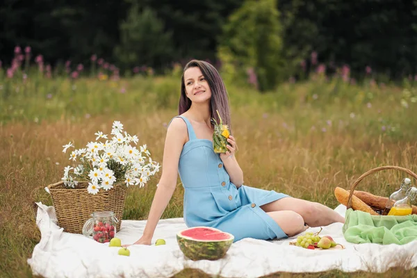 Mladá krásná žena v šatech pije limonádu ze skleněného džbánu a sedí na plání zelené trávy. Piknikový koš, kytice sedmikráků, meloun. — Stock fotografie