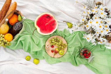 Yeşil çim üzerinde çayırda yaz pikniği. Meyve sepeti, meyve suyu ve şişe şarap, karpuz, çilek bir cam kavanoz ve ekmek Bagetler. Beyaz masa örtüsü ve bir buket alan papatyalar.