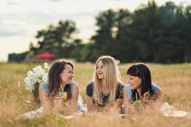 Mavi elbiseler içinde üç genç kadın, ve şapka ekose ve İçecek şarap üzerinde yatıyor. Sahilde çim üzerinde açık piknik. Piknik sepeti ve şarap lezzetli yemek. Karpuz, üzüm ve buket.
