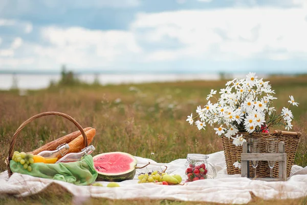 Summer picnic in the meadow on the green grass. Fruit basket, juice and bottled wine, watermelon and bread baguettes. White tablecloth and a bouquet of field daisies.