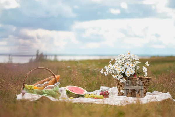 Summer picnic in the meadow on the green grass. Fruit basket, juice and bottled wine, watermelon and bread baguettes. White tablecloth and a bouquet of field daisies.