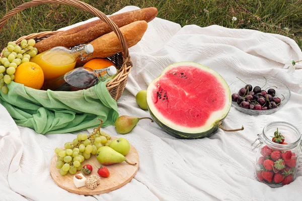 Summer picnic in the meadow on the green grass. Fruit basket, juice and bottled wine, watermelon and bread baguettes. White tablecloth and a bouquet of field daisies.