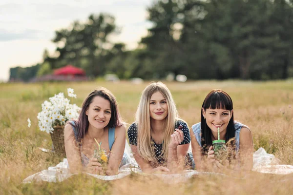 Three young women in blue dresses, and hats lie on plaid and drink wine. Outdoor picnic on grass on beach. Delicious food in picnic basket and wine. Watermelon, grapes and bouquet. — Stock Photo, Image