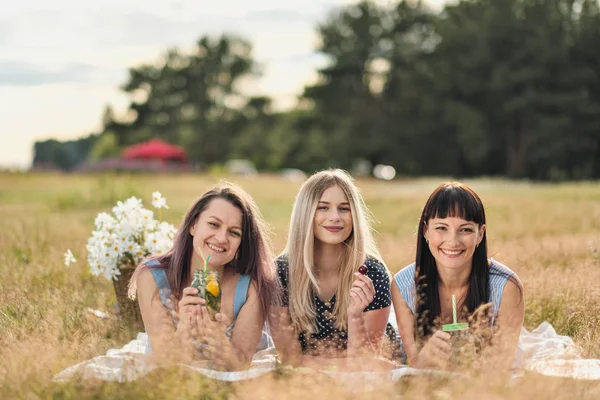 青いドレスを着た3人の若い女性と帽子が格子柄の上に横たわり、ワインを飲みます。ビーチの草の上の屋外ピクニック。ピクニックバスケットとワインの美味しい食べ物。スイカ、ブドウ、ブーケ. — ストック写真