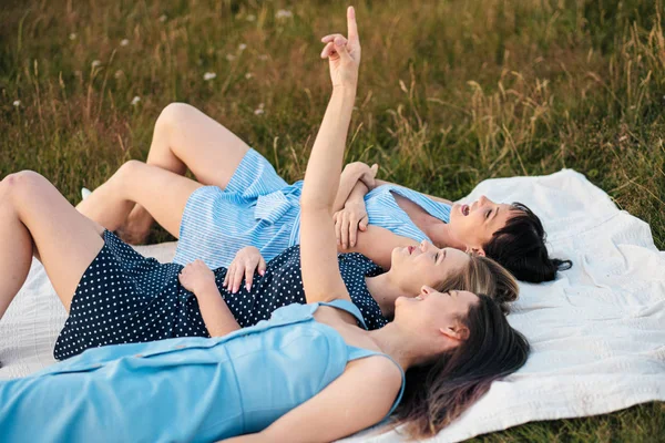 Tre unga attraktiva kvinnor ligger på en pläd och titta på himlen. Skratta och peka upp, dansa. Vila flickvänner utomhus natur. — Stockfoto