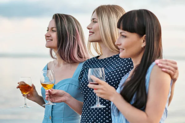 Three young attractive women, in blue dresses are walking along the seashore. Girlfriends communicate, laugh and drink wine.