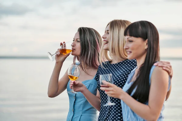 Three young attractive women, in blue dresses are walking along the seashore. Girlfriends communicate, laugh and drink wine.