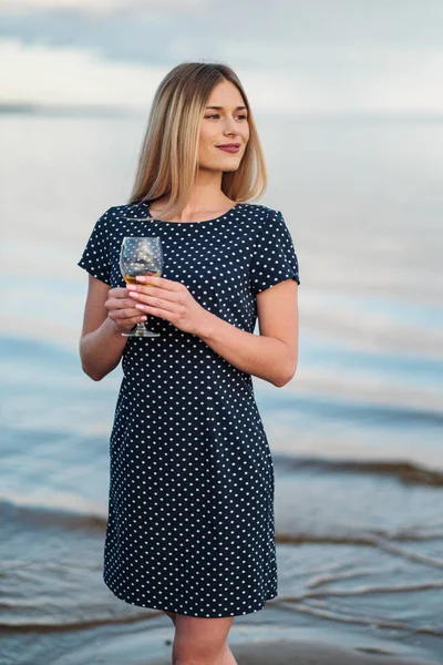 Młoda atrakcyjna blond kobieta, w niebieskiej sukni, spacery wzdłuż brzegu morza. Portret uśmiechniętej kobiety na wakacjach z kielichem wina. — Zdjęcie stockowe