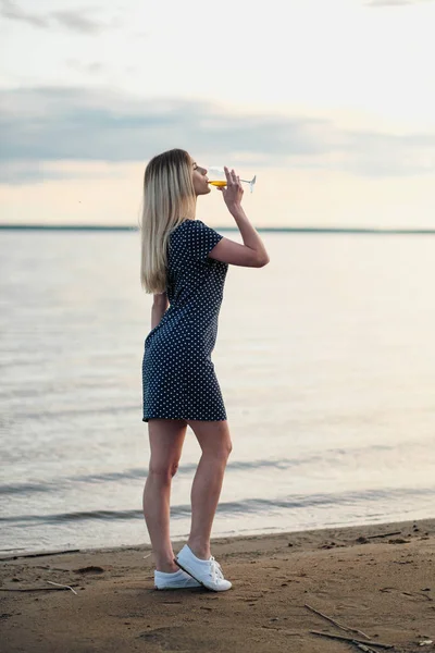 Genç ve çekici bir sarışın kadın, mavi bir elbiseyle, sahil boyunca yürür. Bir bardak şarap ile tatile gülümseyen bir kadının portresi. — Stok fotoğraf