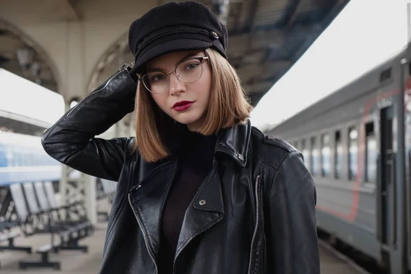 Attractive young woman millenial in black clothes and a hat and glasses at the railway station next to the train. Serious look, waiting for the train.