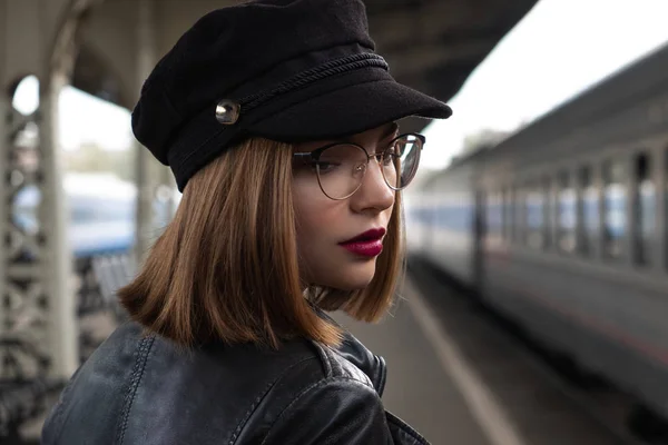 Attractive young woman millenial in black clothes and a hat and glasses at the railway station next to the train. Serious look, waiting for the train.