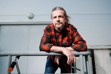 Uzun saçlı yaşlı bir hipster adam ve kırmızı ekose gömlek bir sakal gri bir binaya karşı korkulan üzerinde eğilir.