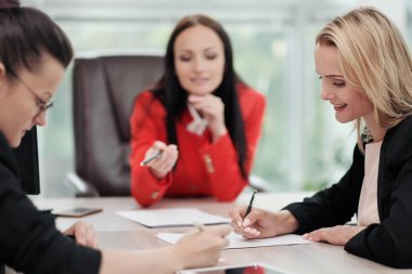 İş elbiseler üç genç çekici kadın bir masada oturuyor ve iş akışları tartışıyor. Baş ve astlar. Profesyonel ve iş arkadaşları çalışma ekibi.