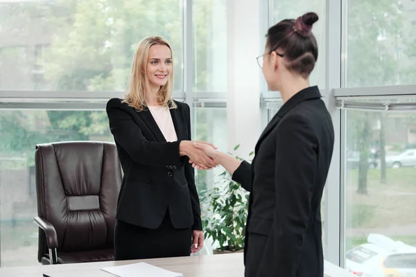 Duas belas mulheres jovens em ternos de negócios apertam as mãos e sorriem. A contratar um emprego. A assinar o acordo. Mulheres no trabalho . — Fotografia de Stock