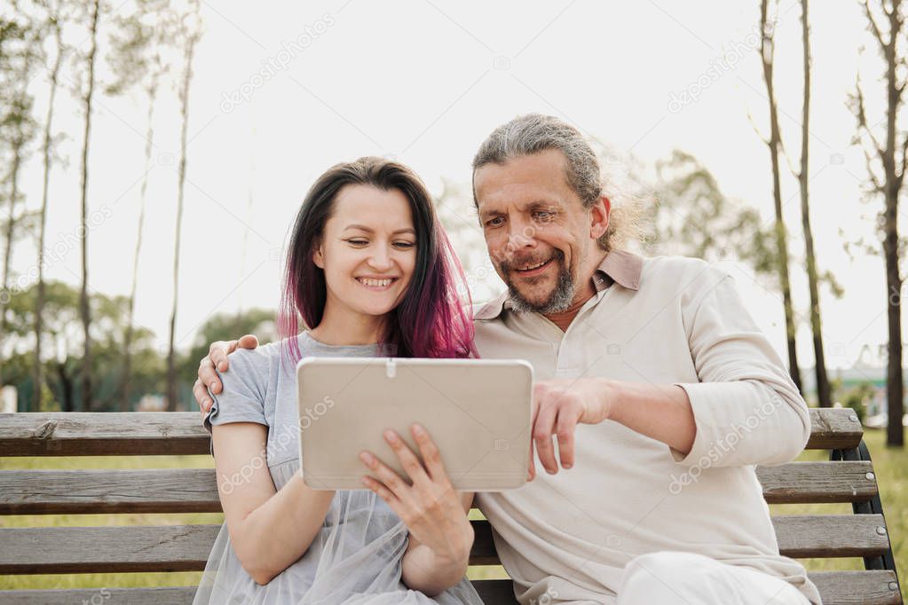 An elderly handsome man with long hair in the tail and a young beautiful woman are sitting on a park bench and using a tablet. Father and daughter relationship.