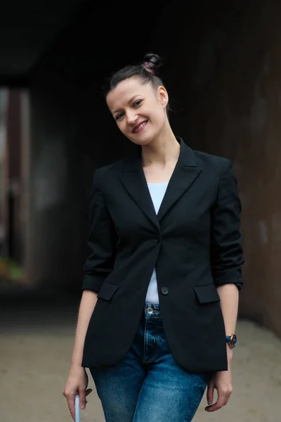 Jovem mulher atraente em casaco preto e jeans azul posando ao ar livre contra o fundo do edifício . — Fotografia de Stock